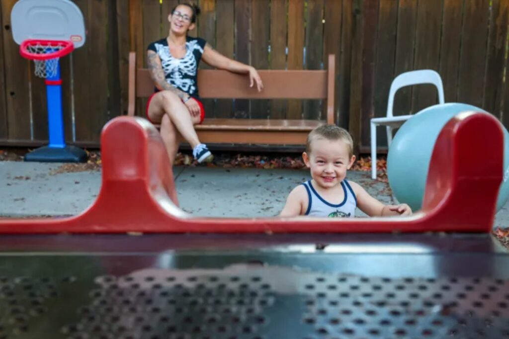 Boy on playground equipment smiles while his mom watches in the background